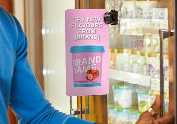 A shopper in the frozen aisle opens a freezer door with an ice cream advertisement next to it.